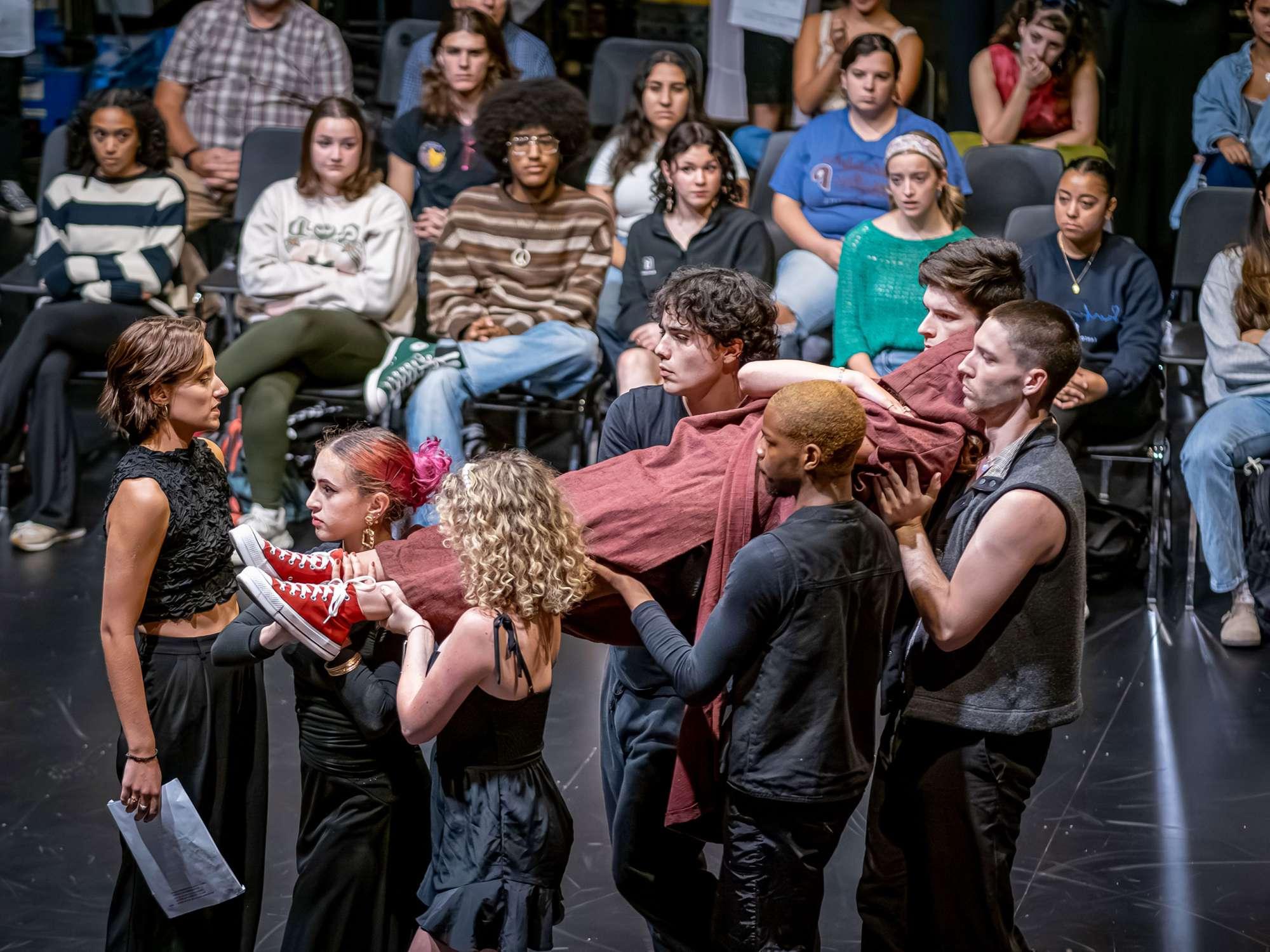 Five actors carry an actor wrapped in cloth as the audience seated on the stage looks on.
