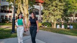 Two female students walking along a campus path with other students sitting on the lawn at Montclair State University.
